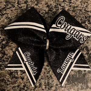 May include: Black and white cheer bow with glitter. The bow has white stripes and the words "Captain", "Lauren", and "Cougar's" written in white.
