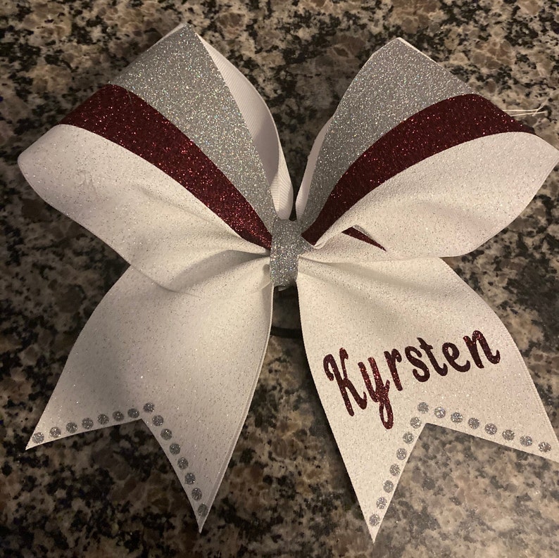 May include: White cheer bow with silver and burgundy stripes and silver glitter. The bow has a silver center and the name "Kyrstem" is written in burgundy glitter on the bottom.