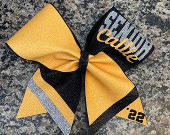May include: A black and gold cheer bow with silver glitter accents. The bow has the text "Senior" and "Callie" in silver glitter on the black portion of the bow. The year "'22" is on the gold portion of the bow.