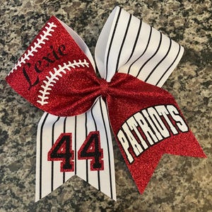 May include: A red glitter baseball bow with the name "Lexie" and baseball stitching. The bow has a white and black striped pattern, with the number "44" and the word "Patriots" in white letters.