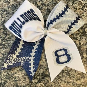May include: A white and navy blue cheerleading bow with glitter accents. The bow features the word "BULLDOGS" in navy blue, the number "8", and the name "Lonaya". The bow has a baseball stitch design.