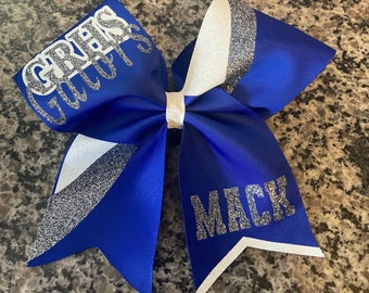 May include: A blue and white cheer bow with silver glitter. The bow has the text "GRAS GIRLS" on one side and "MACK" on the other side.