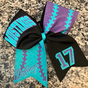 May include: A black cheerleading bow with teal and purple accents. The bow features the word "MUSTANGS" in teal, the name "Destiny" in teal script, and the number "17". The bow has a baseball stitch design.