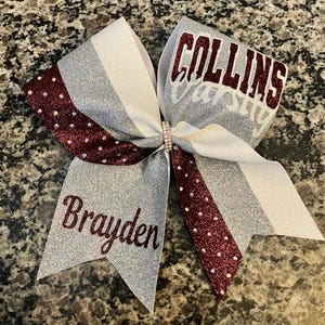May include: A silver and burgundy cheer bow with the words "Collins Varsity" and "Brayden" in burgundy lettering.