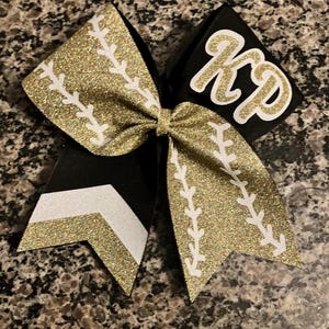 May include: A gold glitter cheerleading bow with white baseball stitching design. The bow has black and white accents and the initials "KP" in white script on a black background. The bow is likely for a sports team.