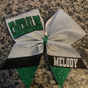 May include: A white, silver, black, and green cheer bow with the words "Cheeralls" and "Melody" in black glitter.