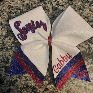 Pu&ograve; includere: Un fiocco da cheerleader bianco glitterato con dettagli rossi e blu. Il fiocco presenta la scritta "Senior" in glitter rosso e blu da un lato e il nome "Gabby" e il numero "23" dall'altro.