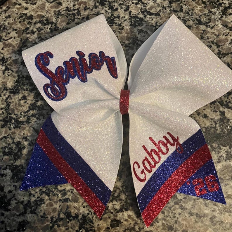 Pu&ograve; includere: Un fiocco da cheerleader bianco glitterato con dettagli rossi e blu. Il fiocco presenta la scritta "Senior" in glitter rosso e blu da un lato e il nome "Gabby" e il numero "23" dall'altro.