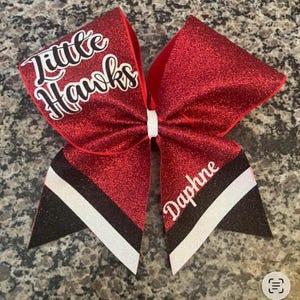 Pu&ograve; includere: Un fiocco da cheerleader rosso glitterato con la scritta "Little Hawks" in corsivo bianco. Il fiocco ha accenti a righe bianche e nere e il nome "Daphne" in corsivo bianco. Il fiocco &egrave; legato con un nastro bianco.