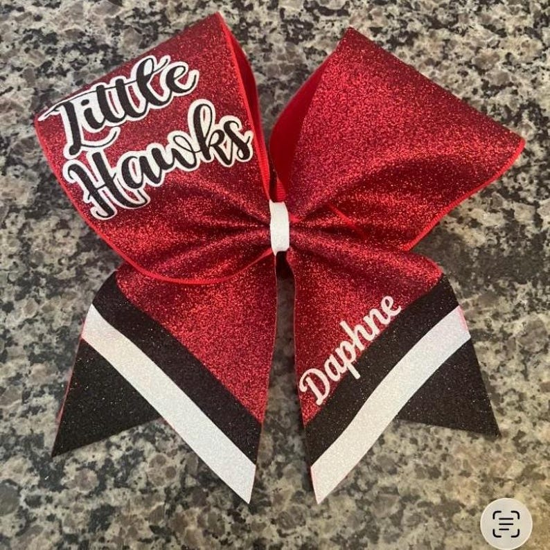 Pu&ograve; includere: Un fiocco da cheerleader rosso glitterato con la scritta "Little Hawks" in corsivo bianco. Il fiocco ha accenti a righe bianche e nere e il nome "Daphne" in corsivo bianco. Il fiocco &egrave; legato con un nastro bianco.