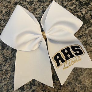 May include: A white cheerleading bow with a gold glitter center. The bow features the letters "AHS" in black with a gold glitter outline and the word "Love" in gold glitter.