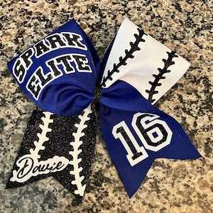 May include: A blue and black cheer bow with white stitching and glitter. The bow has the text "Spark Elite" and the number "16" on it. The bow also has the name "Dawnie" on it.