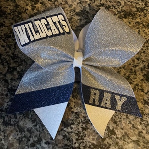 May include: A silver glitter cheer bow with a white and navy blue stripe. The bow has the words "WILDCATS" and "RAY" written on it.