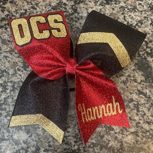 May include: A red and black cheer bow with gold glitter accents. The bow has the text "OCS" in gold letters on one side and the name "Hannah" in gold letters on the other side.