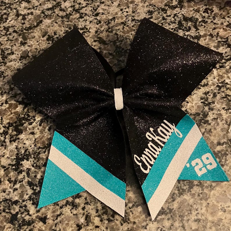 Pu&ograve; includere: Fiocco da cheerleader nero glitterato con dettagli turchese e bianchi. Presenta il nome "Enna Kay" e il numero "'29" in corsivo bianco. Un accessorio decorativo per cheerleader.