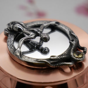 May include: A silver-toned metal pendant featuring a figure of a person reaching out from a circular frame with a snake design. The pendant is on a copper-colored surface.