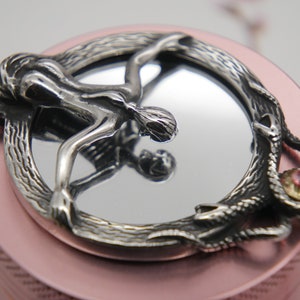 May include: A silver pendant featuring a woman's figure with a snake wrapped around her, holding a mirror with a pink stone. The pendant is set in a circular frame with a textured design.