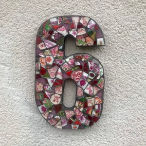 May include: A mosaic number six made of broken pieces of colorful ceramic tiles. The tiles are arranged in a random pattern and include shades of pink, red, and green.