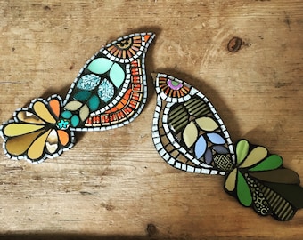Könnte beinhalten: Zwei bunte Mosaik-Vogel-Ornamente mit geometrischen Mustern in Orange-, Grün-, Blau- und Gelbtönen. Die Vögel sind aus kleinen Glas- und Fliesenstückchen gefertigt.