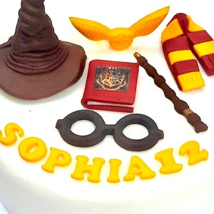 May include: A white cake decorated with a brown wizard's hat, a yellow golden snitch, a red book with the Hogwarts crest, a brown wand, a red and yellow scarf, black round glasses, and the name "Sophia" in yellow icing.