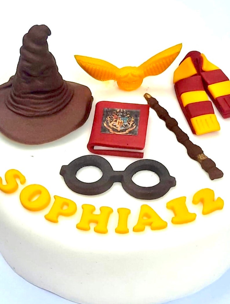 May include: A white cake decorated with a brown wizard's hat, a yellow golden snitch, a red book with the Hogwarts crest, a brown wand, a red and yellow scarf, black round glasses, and the name "Sophia" in yellow icing.