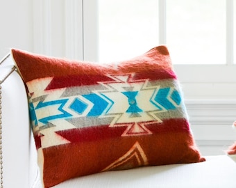 May include: Three decorative throw pillows with Southwestern-inspired designs. The pillows feature geometric patterns in shades of red, white, blue, and gray. They are arranged on a white surface near a window.