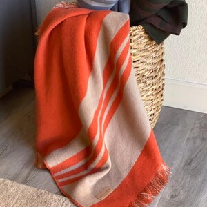 May include: An orange and tan striped blanket with fringe is draped over a wicker basket. Rolled blankets in blue and green are also in the basket. The blanket is on a light-colored rug and a gray wood floor.
