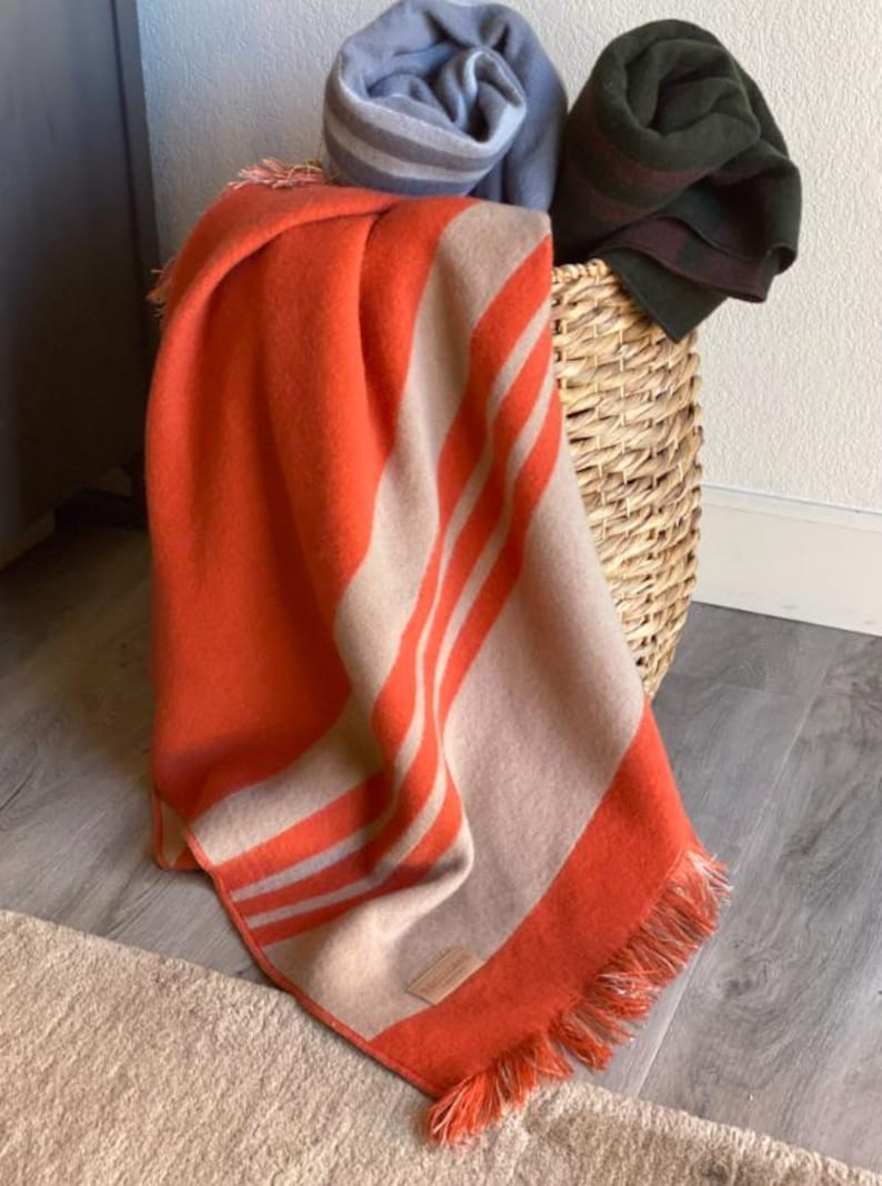 May include: An orange and tan striped blanket with fringe is draped over a wicker basket. Rolled blankets in blue and green are also in the basket. The blanket is on a light-colored rug and a gray wood floor.