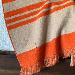 May include: An orange and beige striped blanket with fringe. The blanket has thick orange stripes alternating with beige stripes. The blanket is folded over a surface, with the brand name "DECUADANE" visible on a small tag.