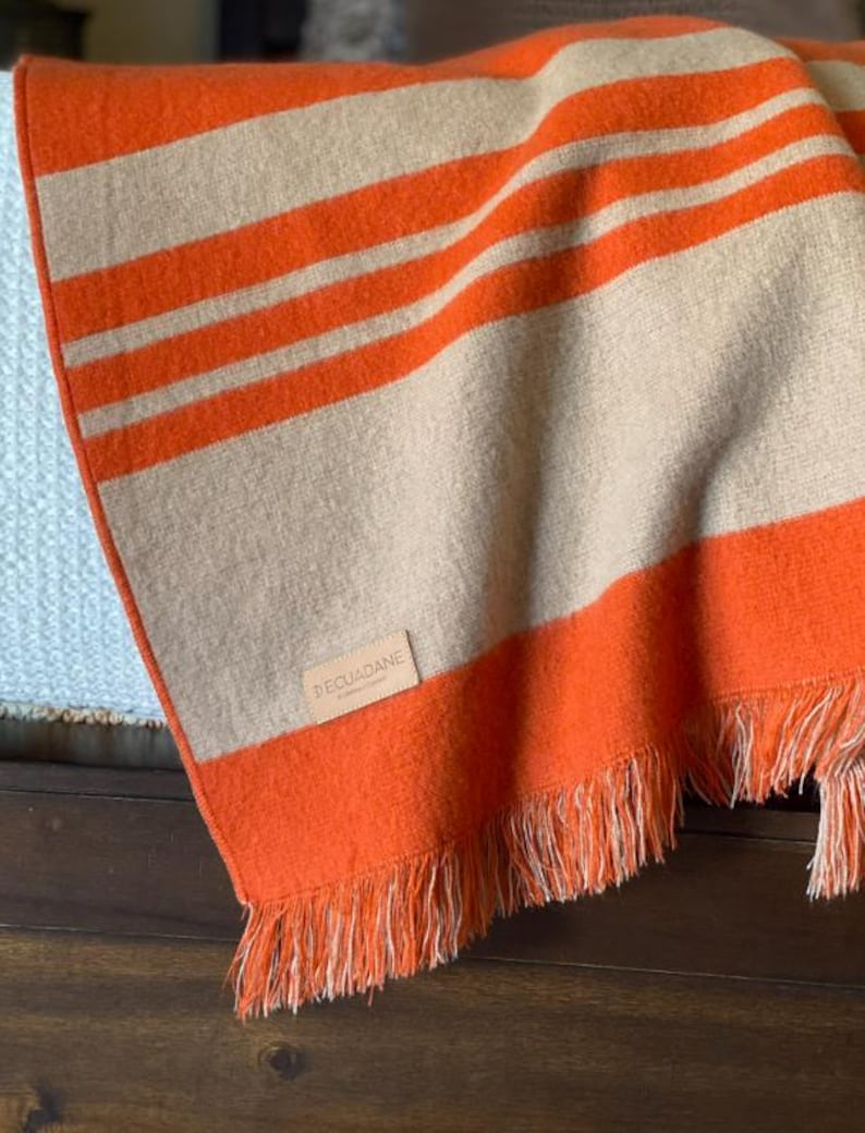 May include: An orange and beige striped blanket with fringe. The blanket has thick orange stripes alternating with beige stripes. The blanket is folded over a surface, with the brand name "DECUADANE" visible on a small tag.
