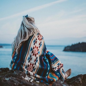May include: A woman sits on a rocky shore wearing a blue and white patterned blanket with red and yellow accents. The blanket is wrapped around her and she is looking out at the ocean.