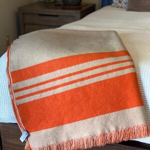 May include: A folded blanket with a tan base and bold orange stripes. The blanket has a fringed edge and is draped over a wooden bed frame. The blanket is a cozy home decor item.