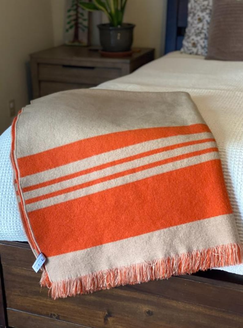 May include: A folded blanket with a tan base and bold orange stripes. The blanket has a fringed edge and is draped over a wooden bed frame. The blanket is a cozy home decor item.