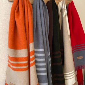 May include: A collection of colorful blankets hanging on a wooden rack. The blankets are in shades of orange, gray, dark green, beige, and red. Each blanket has a striped design and fringed edges. The blankets are made of a soft material.