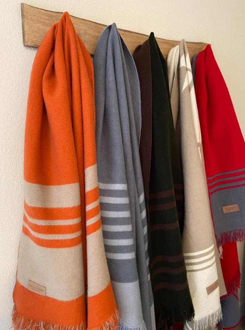 May include: A collection of colorful blankets hanging on a wooden rack. The blankets are in shades of orange, gray, dark green, beige, and red. Each blanket has a striped design and fringed edges. The blankets are made of a soft material.