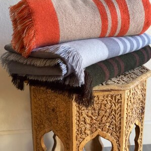 May include: A stack of four folded blankets with fringed edges. The top blanket is orange and tan with orange stripes. The other blankets are gray, gray with blue stripes, and dark green with maroon stripes. The blankets are on a carved wooden table.