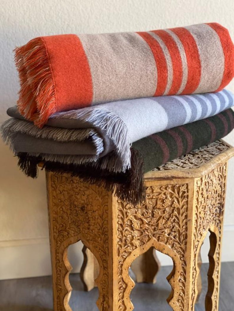 May include: A stack of four folded blankets with fringed edges. The top blanket is orange and tan with orange stripes. The other blankets are gray, gray with blue stripes, and dark green with maroon stripes. The blankets are on a carved wooden table.