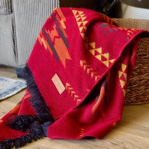 Peut inclure: Une couverture rouge avec un motif géométrique en jaune, orange et noir. La couverture a des franges noires et une étiquette en cuir marron avec le mot "Wanderlust" dessus.