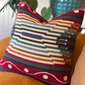 May include: A colorful throw pillow with a geometric pattern in shades of red, blue, green, brown, and white. The pillow has a woven texture and a fringed edge.