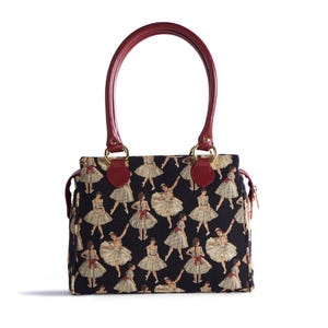 May include: A black handbag with a repeating pattern of ballerinas in cream-colored tutus and red bows. The bag has a burgundy handle and accents, with gold-colored hardware. The bag is rectangular with a zipper closure.