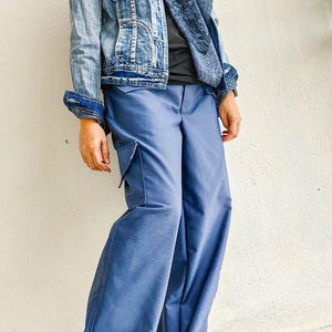 May include: A pair of wide-leg, blue denim cargo pants with a large cargo pocket on the left leg. The pants are cuffed at the bottom and are worn with a denim jacket and a black shirt.