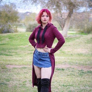 May include: A woman with pink hair is wearing a maroon long-sleeved hoodie with a black drawstring and a high-low hemline. She is also wearing a denim mini skirt and black thigh-high boots.