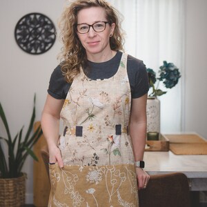 May include: A woman wearing a floral print apron with a large pocket. The apron has a brown and white floral pattern on the top and a solid brown with white floral pattern on the bottom. The apron has a black strap across the waist.