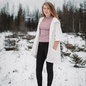 May include: A woman wearing a white cardigan over a pink and purple long-sleeved shirt, black leggings, and black boots. She is standing in a snowy field with trees in the background.