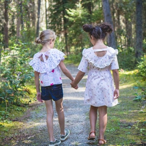 May include: Two girls wearing floral dresses walk hand-in-hand on a path through a forest. The girl in the front is wearing a pink and white dress with a frilly collar and a white bow at the back. The girl in the back is wearing a pink and white dress with a frilly collar and a white lace trim.