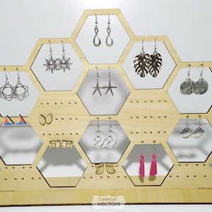 May include: Wooden hexagonal earring stand with a variety of earrings displayed. The stand has a triangular base and is made of light wood. The earrings are made of metal and feature various designs, including stars, moons, suns, leaves, and seashells. The stand is labeled "Lasercut vectors".
