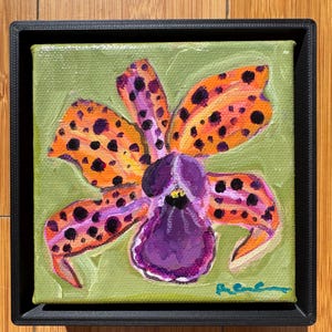 May include: A framed painting of a vibrant orchid flower. The petals are orange with black spots, and the center is purple. The background is a light green, and the painting is in a black frame. The artwork is signed in the lower right corner.