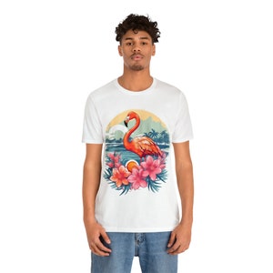 May include: White t-shirt with a colorful graphic of a flamingo standing in water with tropical flowers and palm trees. The graphic is in a circle with a sunset background.