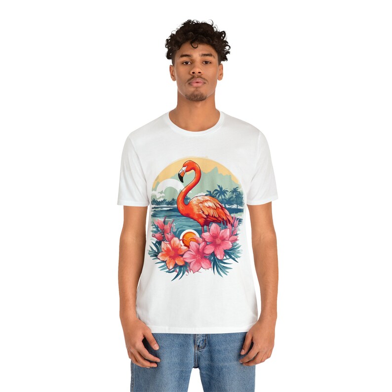 May include: White t-shirt with a colorful graphic of a flamingo standing in water with tropical flowers and palm trees. The graphic is in a circle with a sunset background.