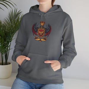 May include: A gray hooded sweatshirt featuring a detailed crest design. The crest includes a golden crown, a red and black eagle, and a shield with a central emblem. The text "CARPE DIEM" is displayed on a banner below the shield.